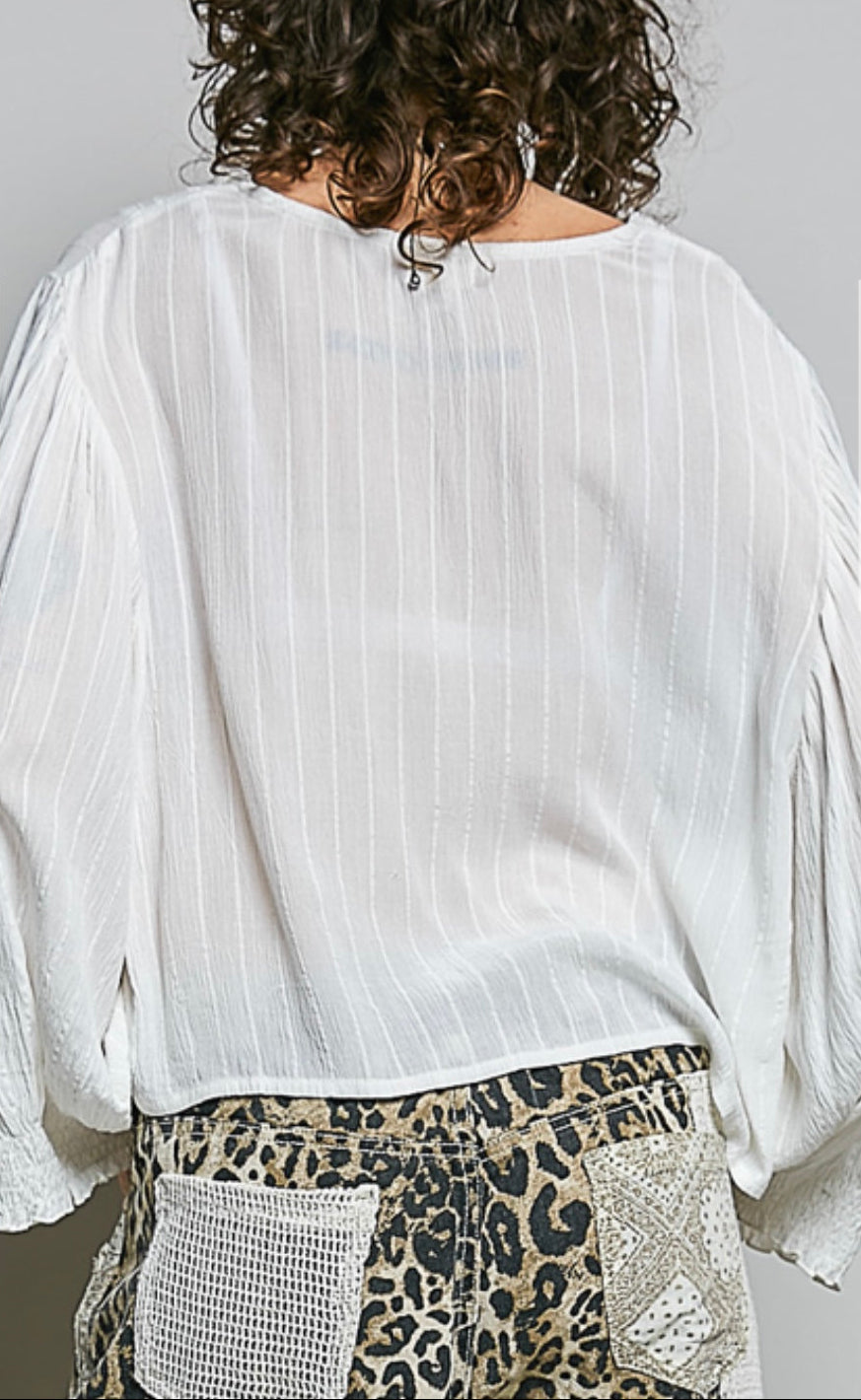 A person shown from the back wearing a loose white long-sleeve blouse with subtle vertical texture and gathered sleeves, paired with leopard-print bottoms. The photo is cropped from the shoulders to the hips against a plain light gray background.