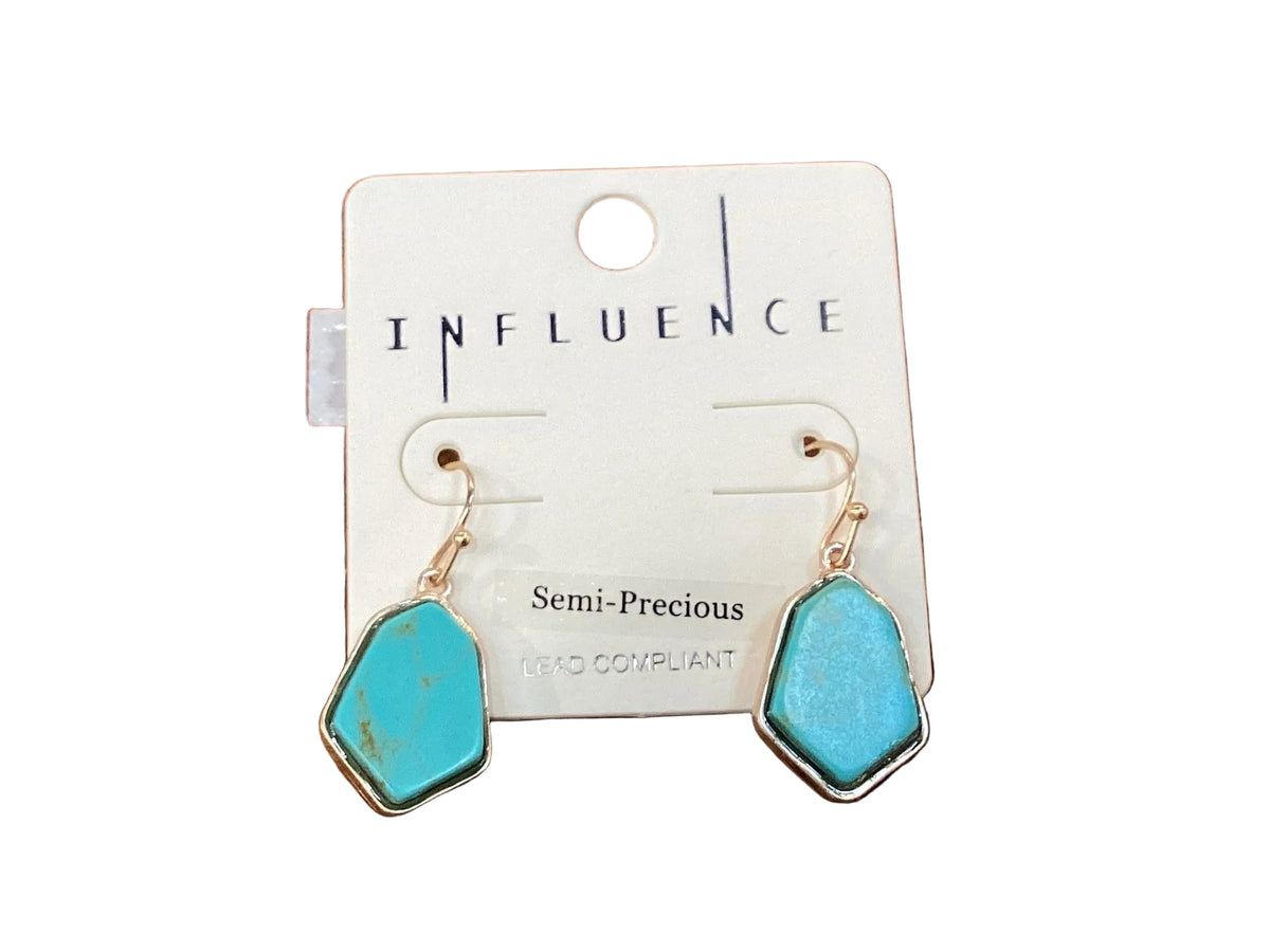 A pair of turquoise semi-precious dangle earrings with gold-tone hooks and silver-tone bezel settings, displayed on a white card labeled 'INFLUENCE' on a wooden surface.