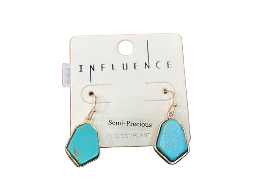 A pair of turquoise semi-precious dangle earrings with gold-tone hooks and silver-tone bezel settings, displayed on a white card labeled 'INFLUENCE' on a wooden surface.