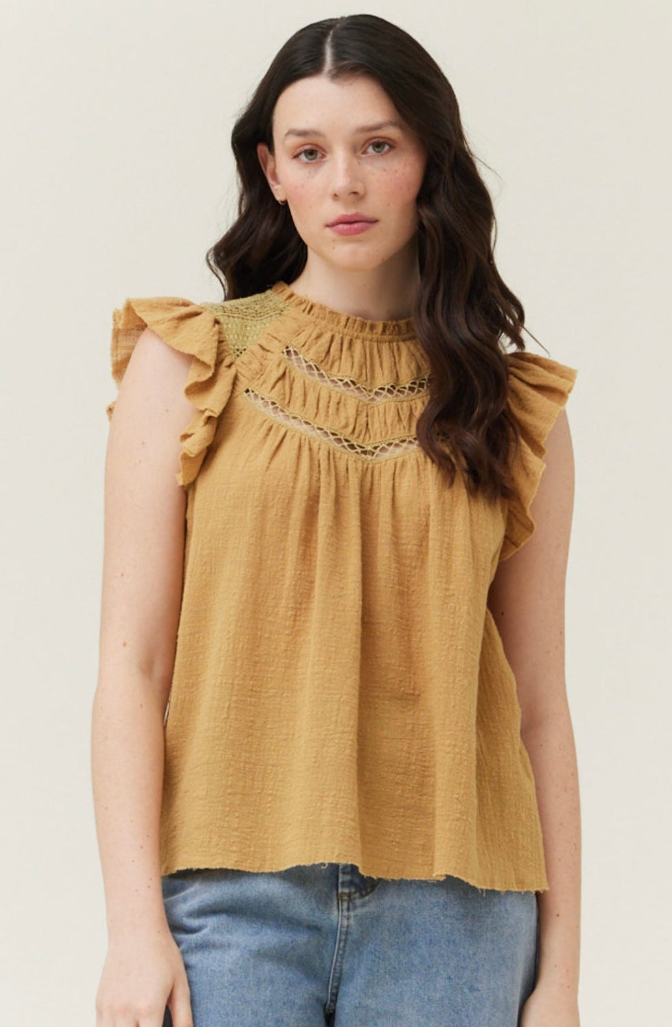 Woman wearing a mustard yellow top with ruffled sleeves and blue jeans on a light background