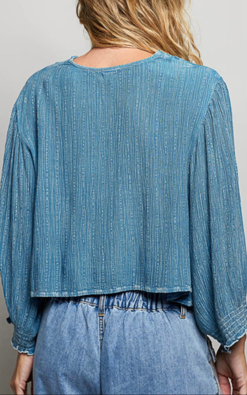 A woman viewed from the back wearing a cropped, long-sleeve blue top with a textured vertical stripe pattern and frayed edges, paired with light blue jeans against a plain light gray background.