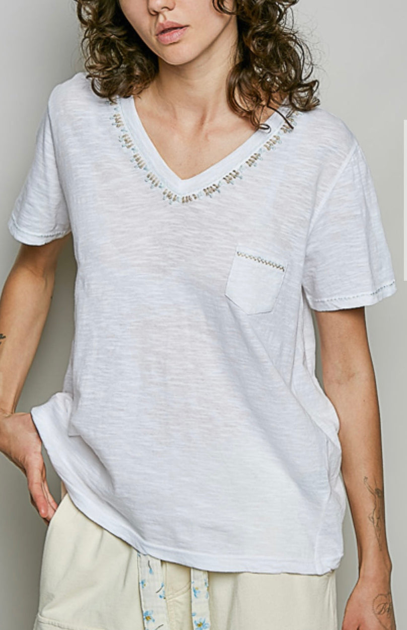 A model with curly dark hair wearing a white short-sleeve V-neck t-shirt with embroidered details around the neckline and a chest pocket, paired with light beige cargo shorts.