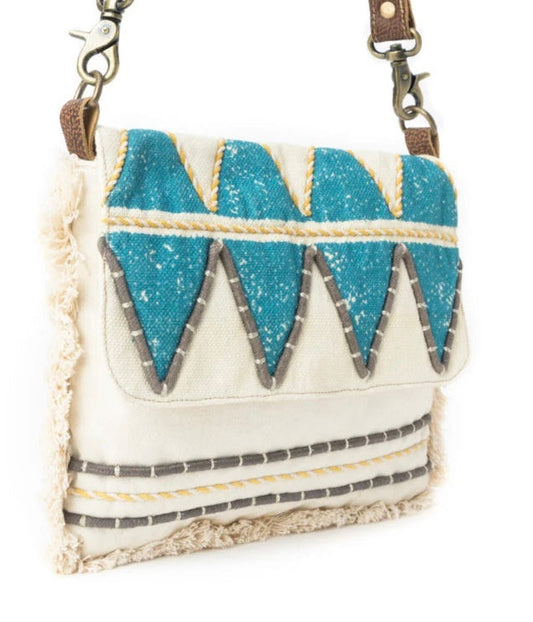 A western-style crossbody bag with a cream canvas body, turquoise blue sequined triangle details, gray and yellow embroidered accents, and fringed edges, shown on a white background.