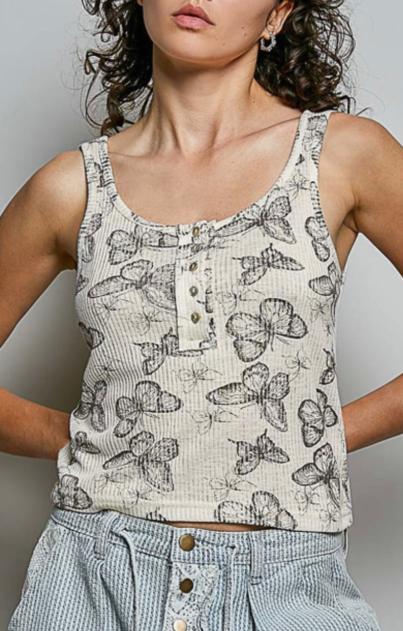 A woman wearing a sleeveless white ribbed tank top with a black butterfly print and a button placket, paired with light blue striped button-up pants, against a plain gray background.