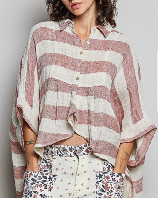 A model wearing a loose, striped linen button-up shirt in dusty rose and cream, paired with cream patchwork trousers featuring floral details, set against a plain gray background.